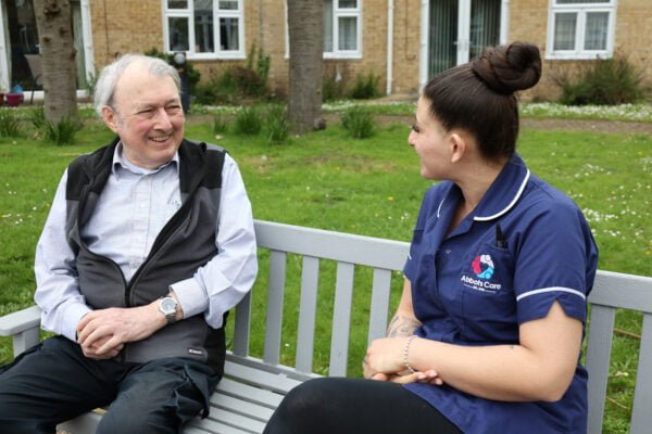 What do carers do: A smiling Abbots Care service user (left) sitting outside on a bench next to an Abbots carer (right).