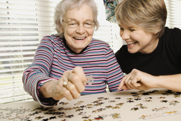 An elderly mother and her daughter laughing together as they complete a puzzle together on the table.