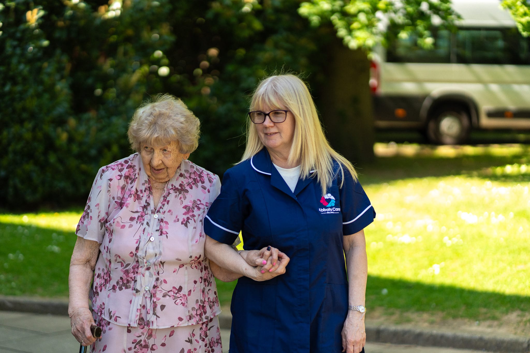 Abbots Care worker walking outdoors with an elderly woman, supporting her with a gentle smile.