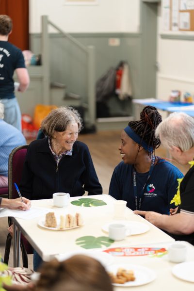An elderly female client laughs with a Care Worker at a client tea party